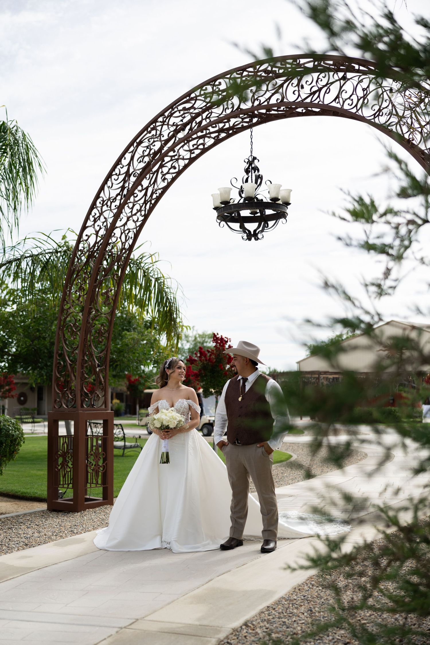 Bride and groom stand under ornate iron arch with chandelier in lush garden wedding venue.