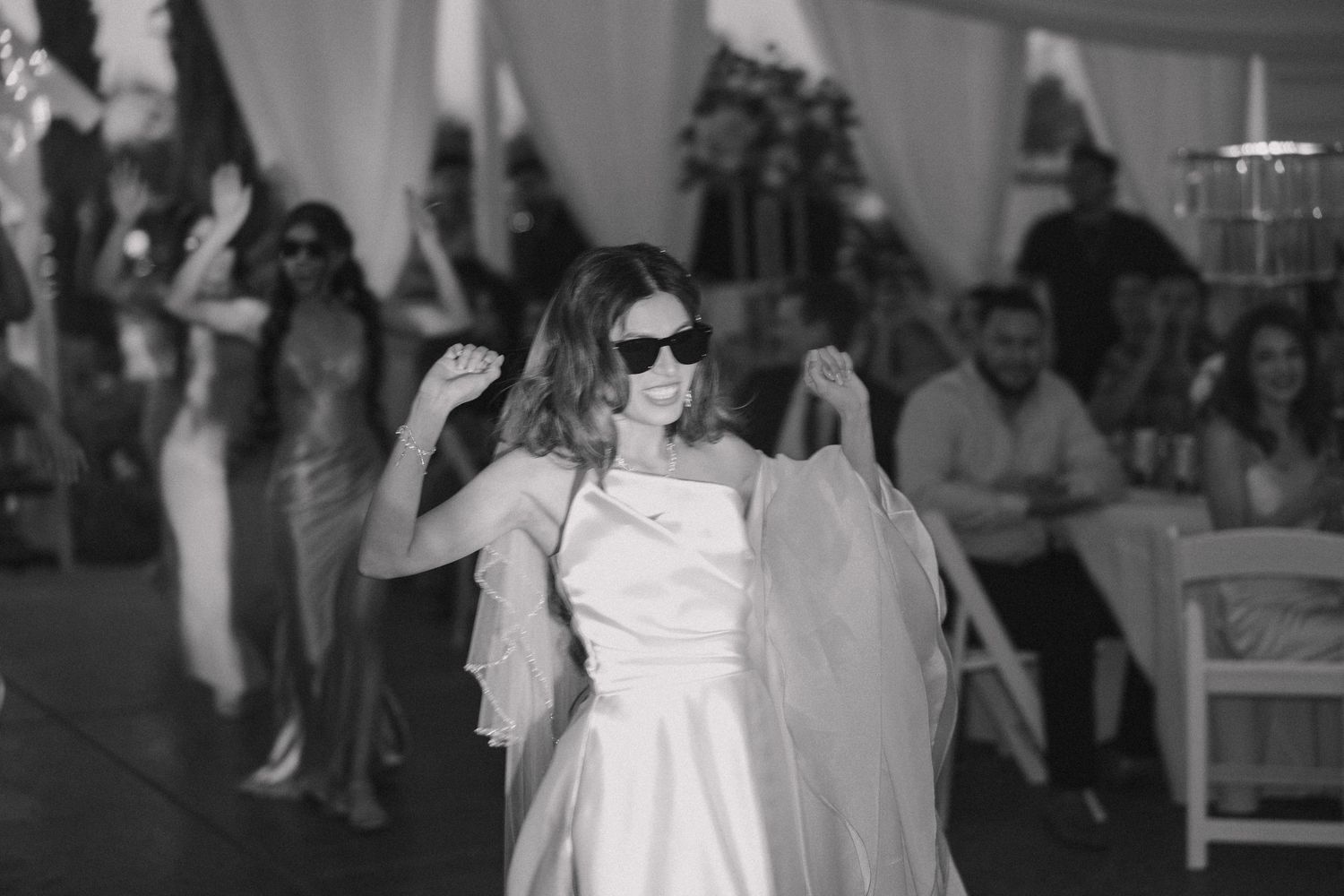 Bride dancing joyfully in sunglasses while holding her veil at a wedding reception.