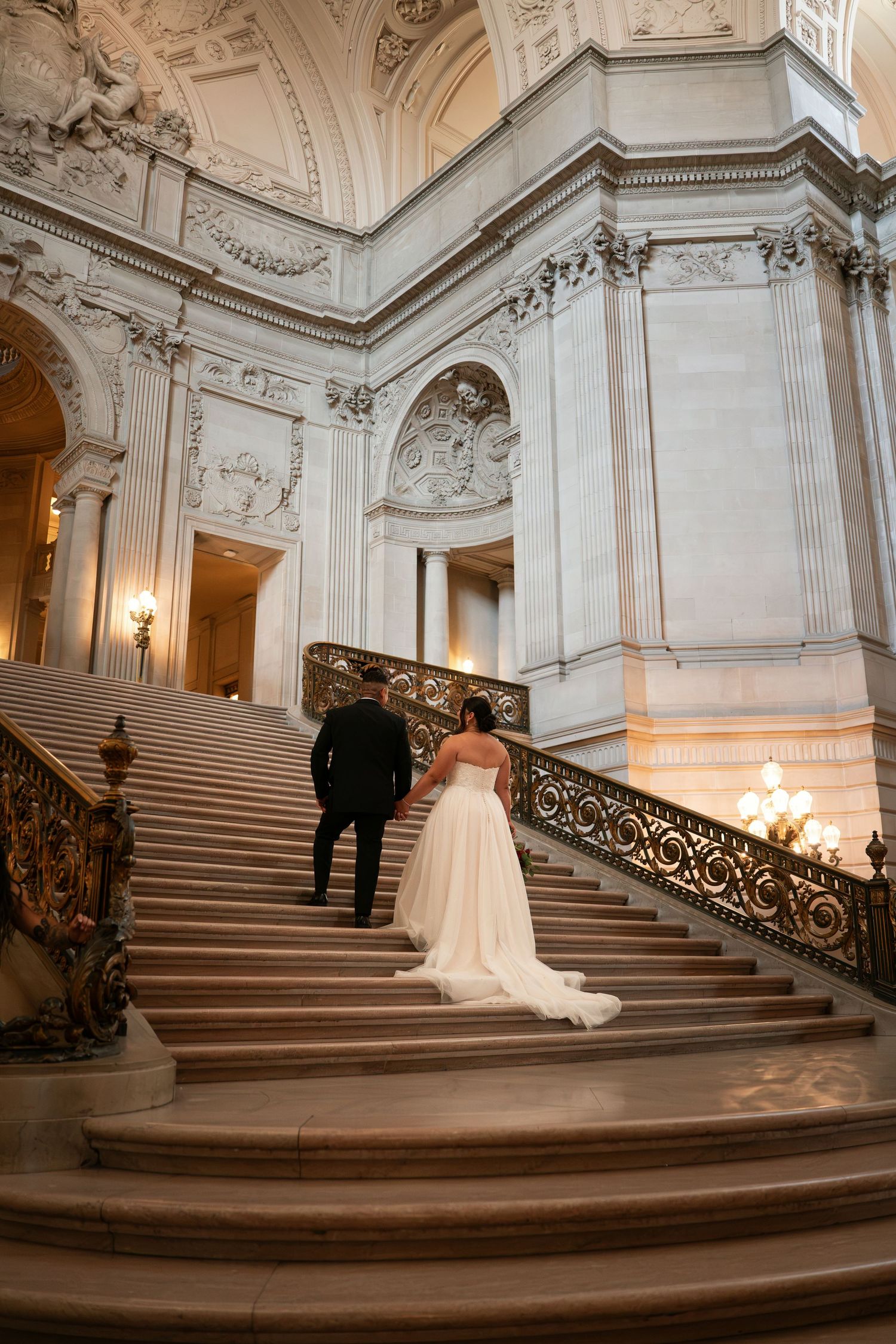 Bride and groom ascending grand marble staircase at San Francisco City Hall, bride in flowing white gown.