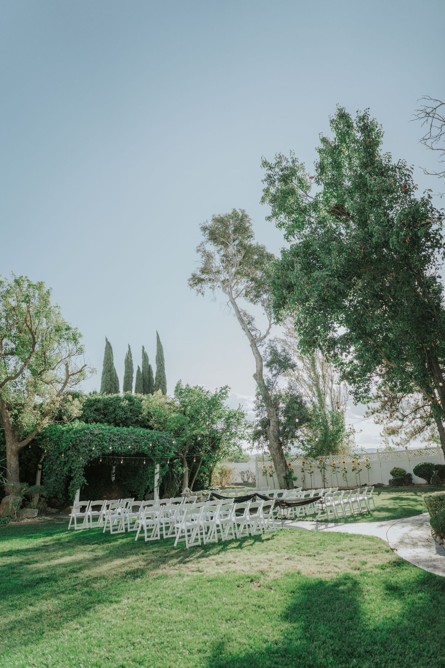 Outdoor wedding ceremony setup with white chairs on green lawn surrounded by lush trees under a clear blue sky.