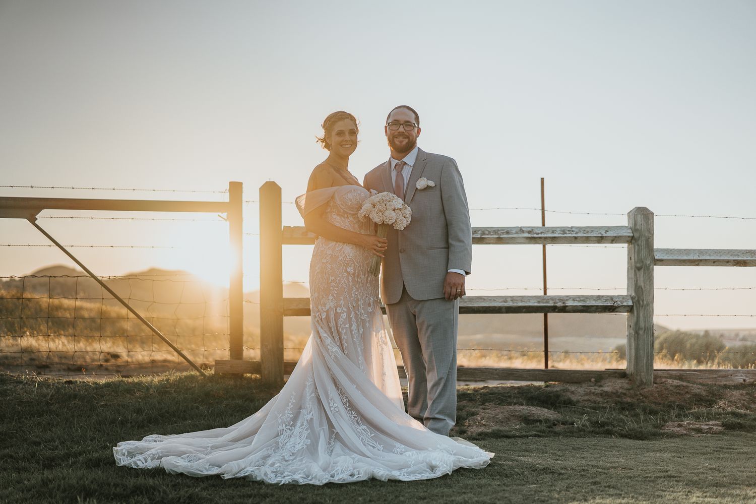 A bride and groom pose at golden sunset by a rustic fence, bride in lace gown, groom in gray suit.