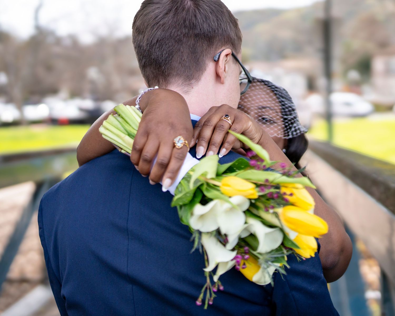 Interracial couple embracing at wedding, bride holding yellow tulips and white calla lilies bouquet, wearing veil.