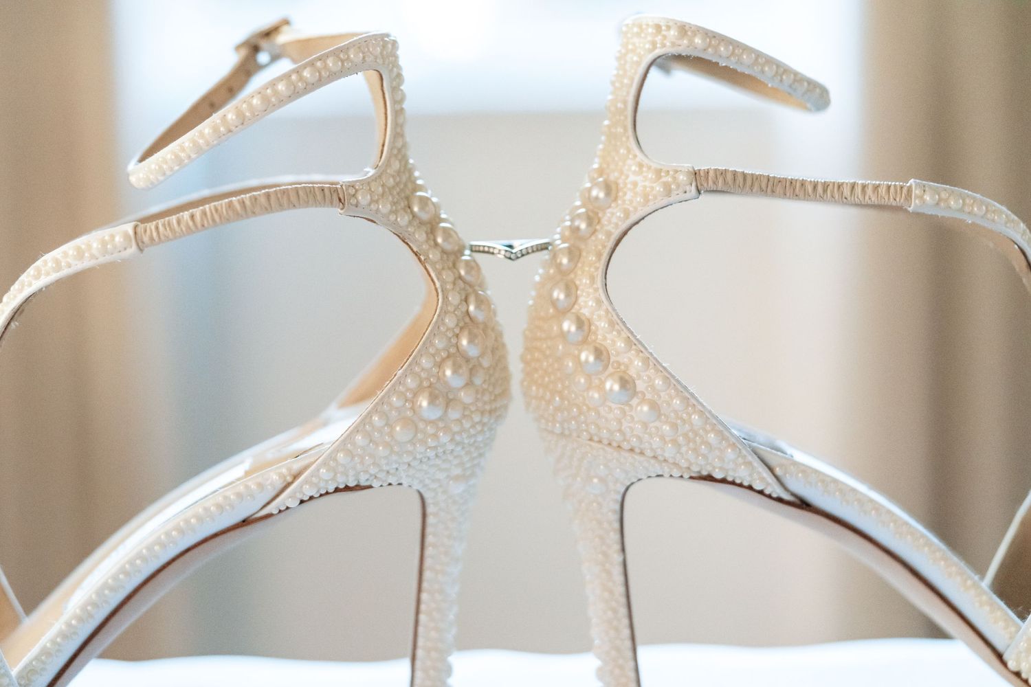 Elegant white bridal heels with pearl embellishments and ankle straps displayed back-to-back on a stand.