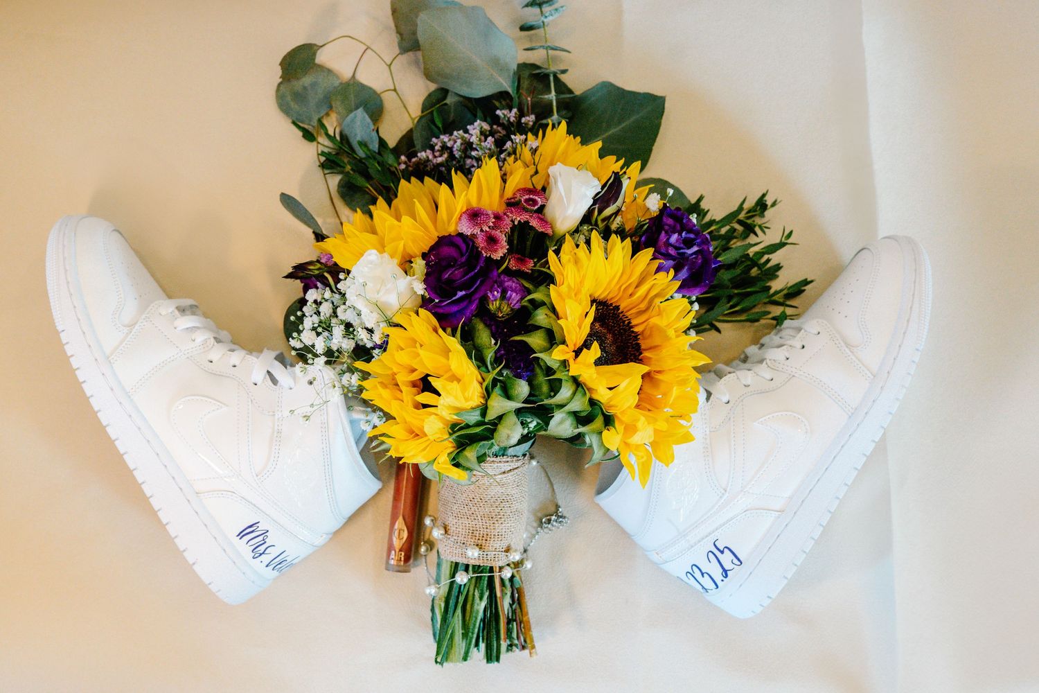 White Nike high-top sneakers beside a vibrant sunflower wedding bouquet with purple flowers on a white background.
