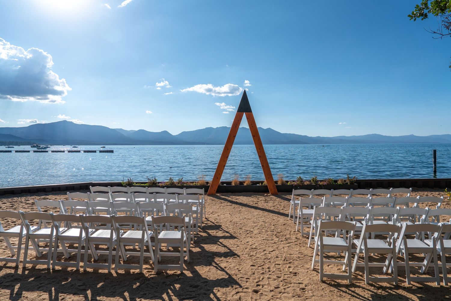 Outdoor lakeside wedding ceremony setup with white chairs, wooden arch, and mountain views under a sunny blue sky.