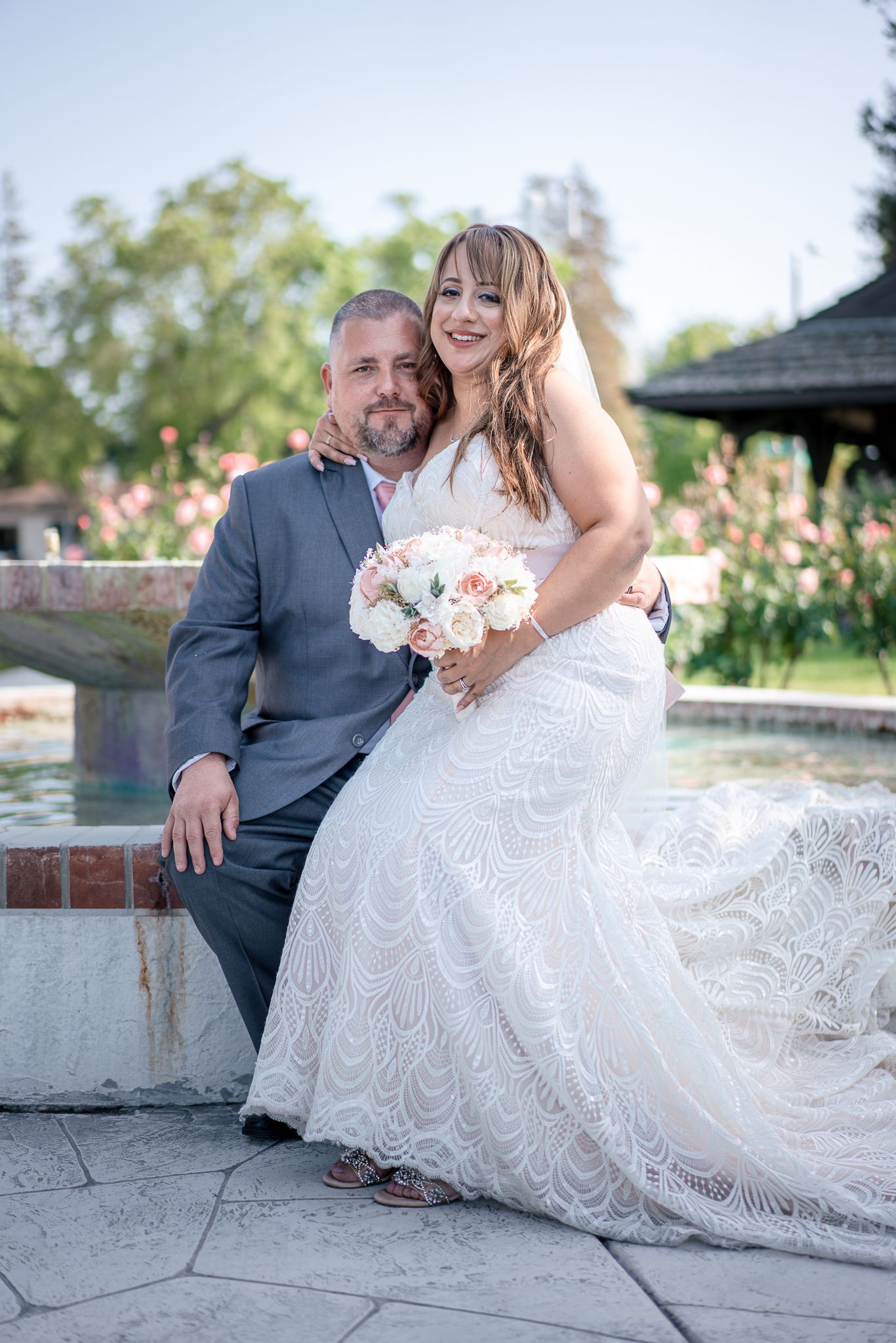 Wedding couple sits together on stone wall in garden setting.