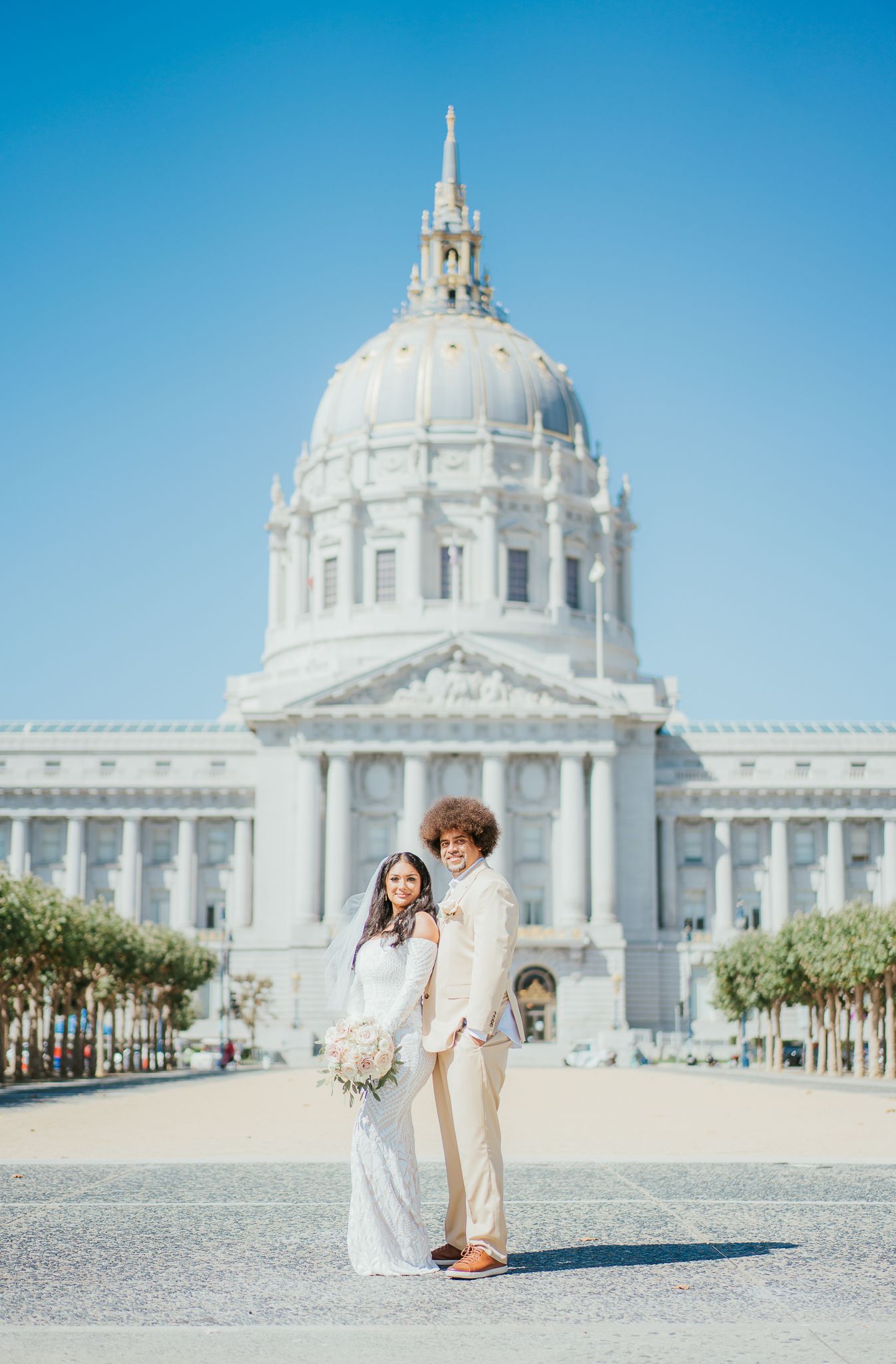 A couple in wedding attire poses in front of San Francisco City Hall's iconic domed architecture on a sunny day.