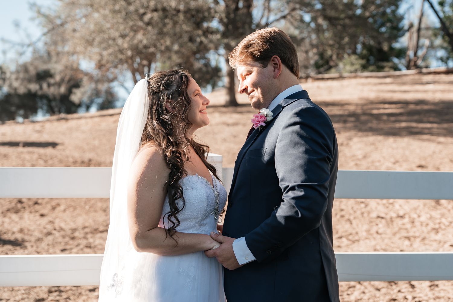 Bride in a white lace gown and veil holds hands with a groom in a navy suit beside a white wooden fence at an outdoor wedding.