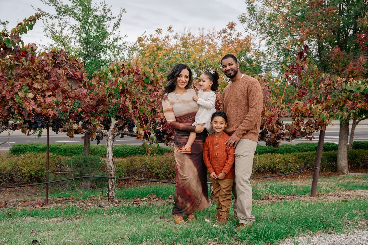 Family portrait session in a park with autumn trees and warm earth-toned clothing.