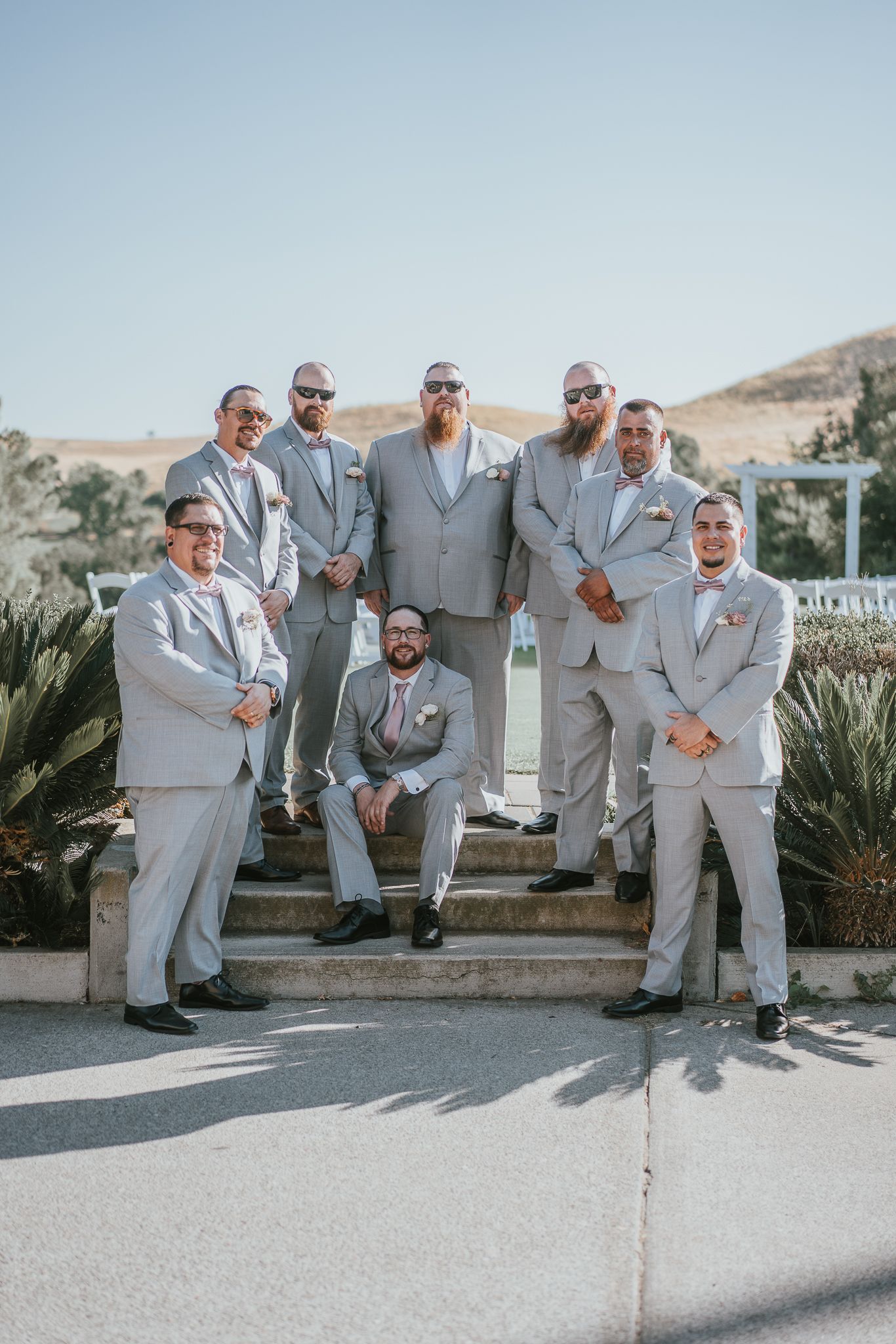 Groomsmen in light gray suits with pink bow ties pose on outdoor steps at a sunny wedding venue.