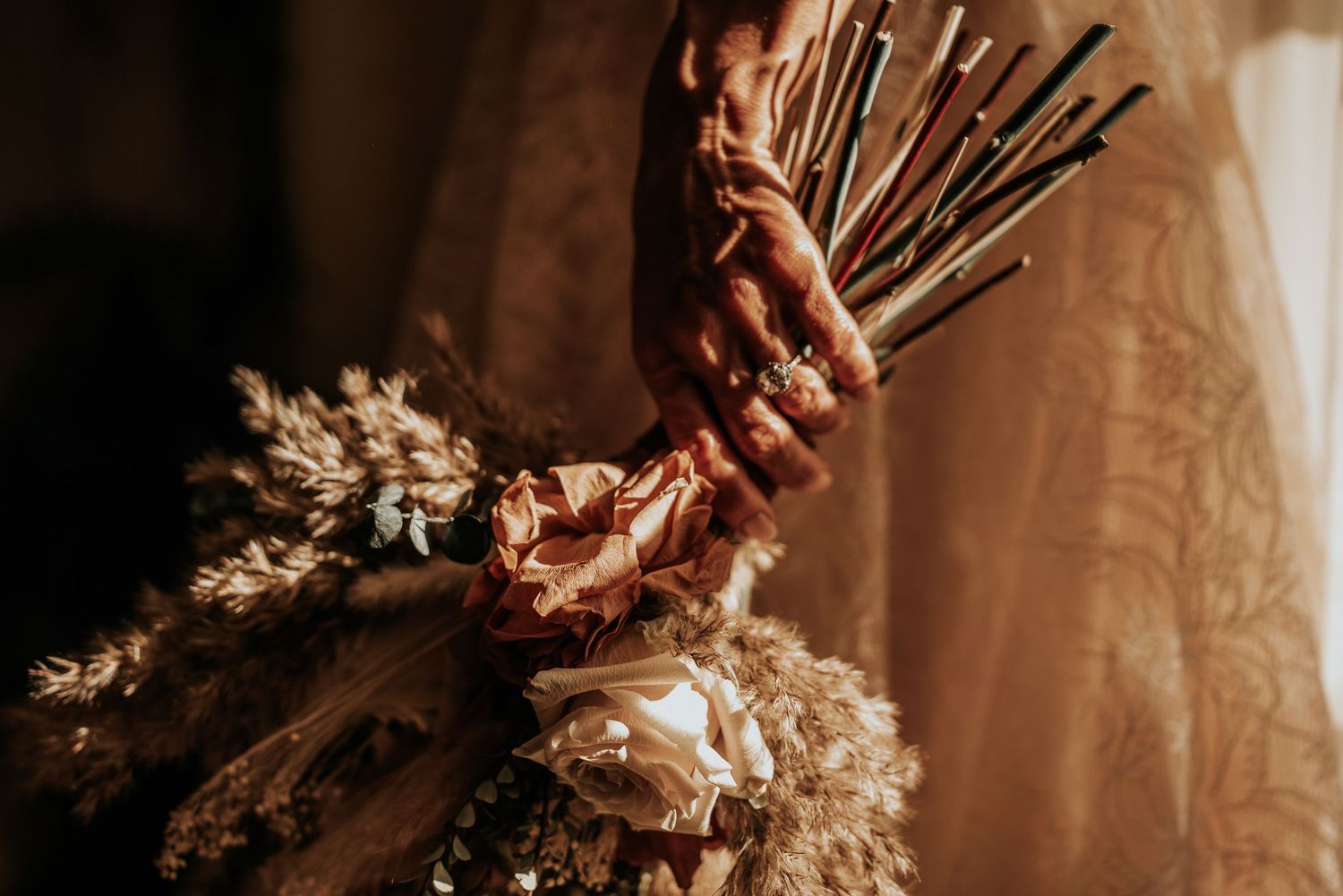Close-up of a bride holding a rustic bouquet of dried flowers and pampas grass with elegant gold rings on her fingers.
