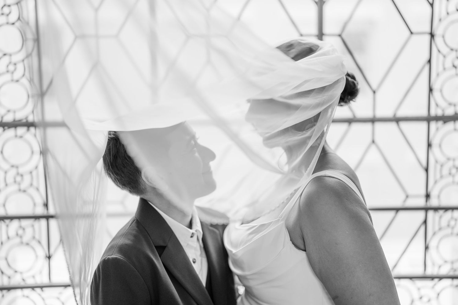 Black and white photo of a couple sharing a romantic moment under a flowing bridal veil at their wedding.