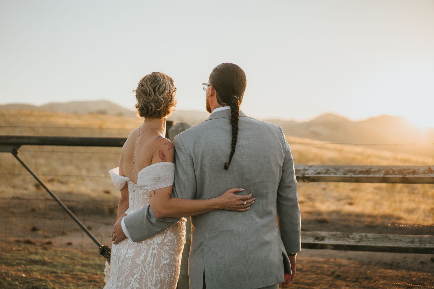 Bride and groom embrace while gazing at golden sunset over rolling hills at outdoor wedding venue.