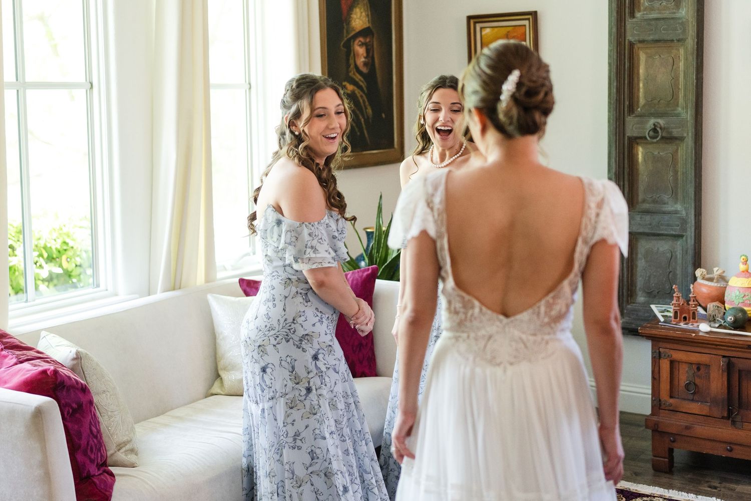 Bride in backless lace gown reveals dress to excited bridesmaids in floral dresses during wedding preparations.