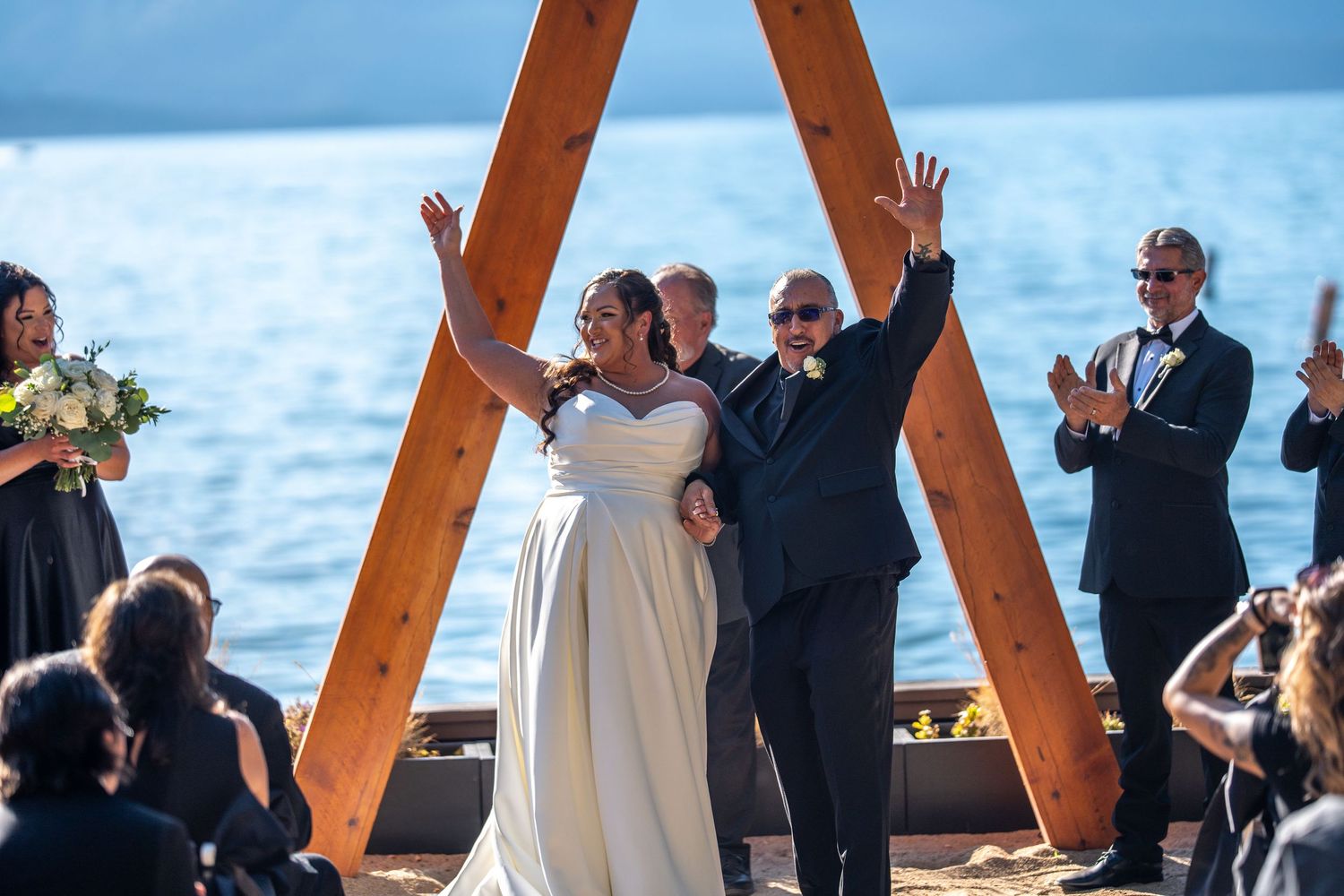 Joyful newlyweds celebrate at outdoor lakeside wedding ceremony under wooden arch, guests applauding in background.