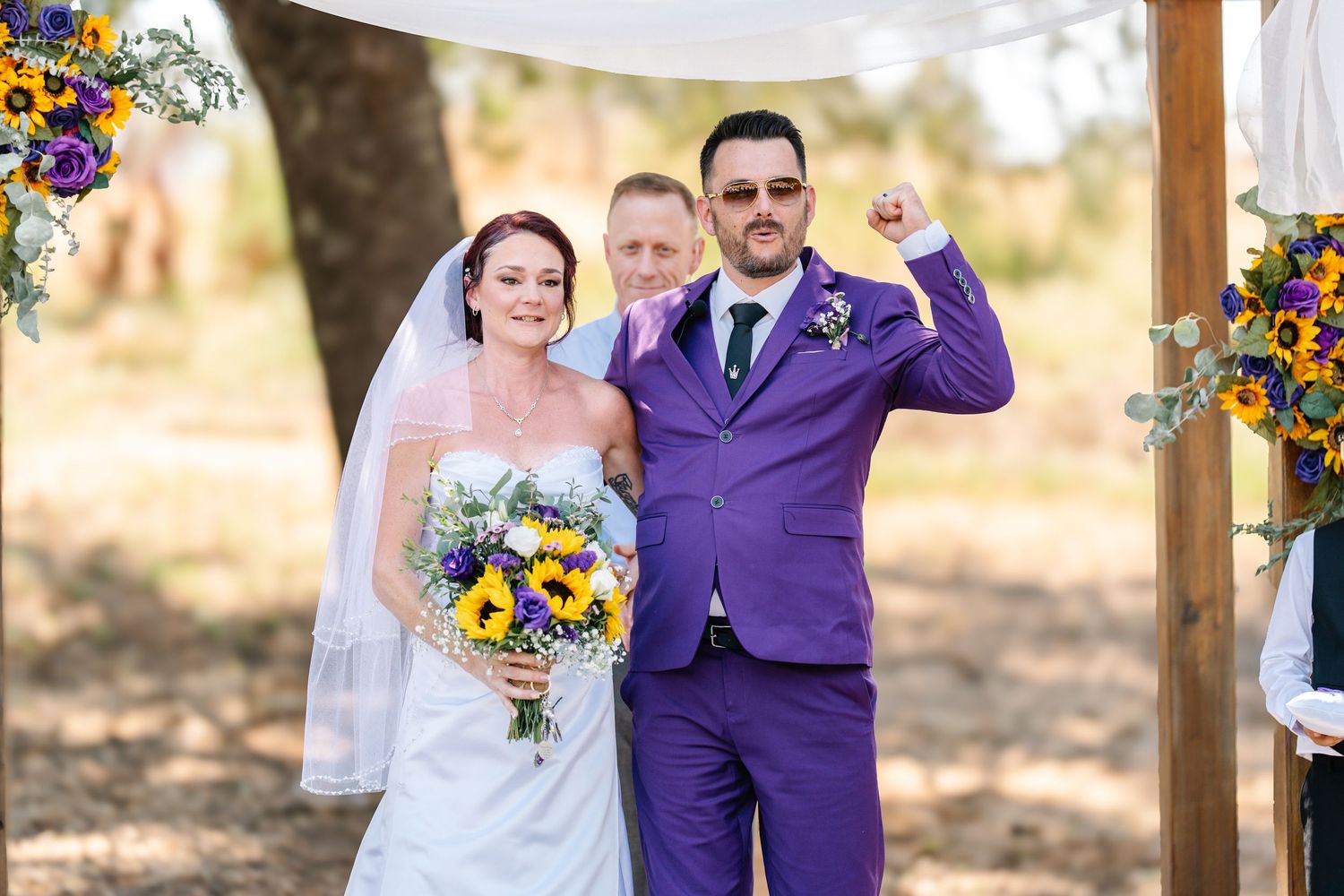 Bride in white gown holding sunflower bouquet stands with groom in purple suit raising fist at outdoor wedding ceremony.