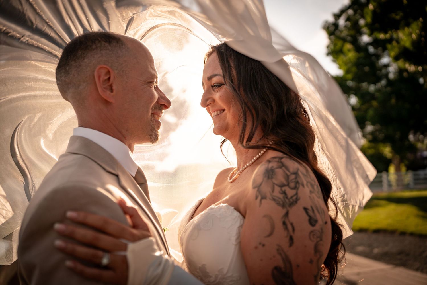 A bride and groom share a romantic moment under a flowing veil, with golden sunset light and floral tattoos visible.