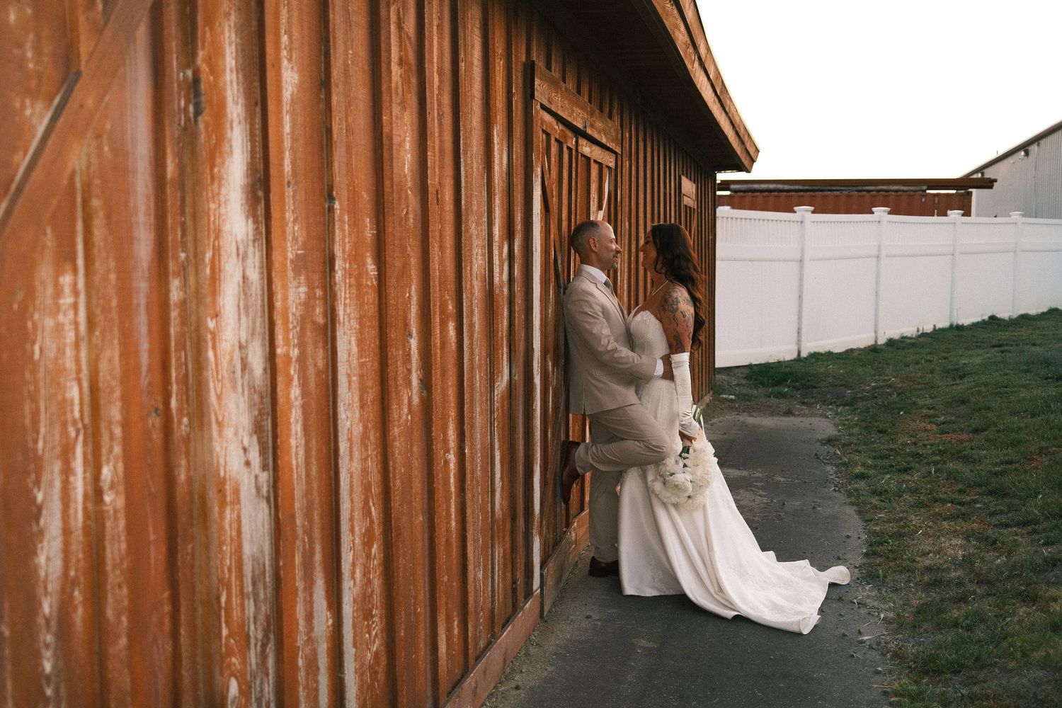 A bride and groom share a romantic moment leaning against a rustic wooden barn wall on their wedding day.