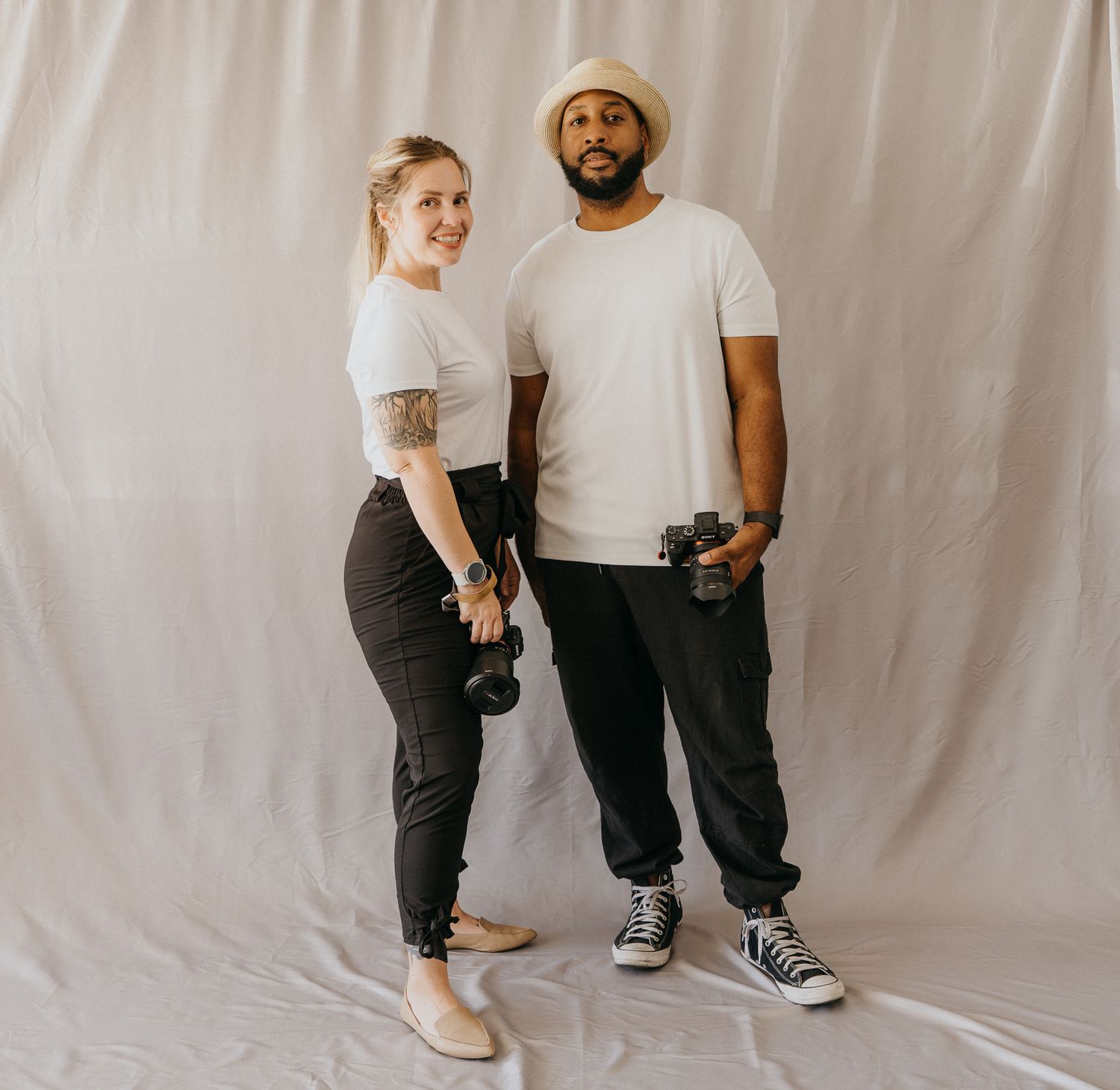 Two photographers in matching white tees and black pants pose with cameras against a white backdrop.