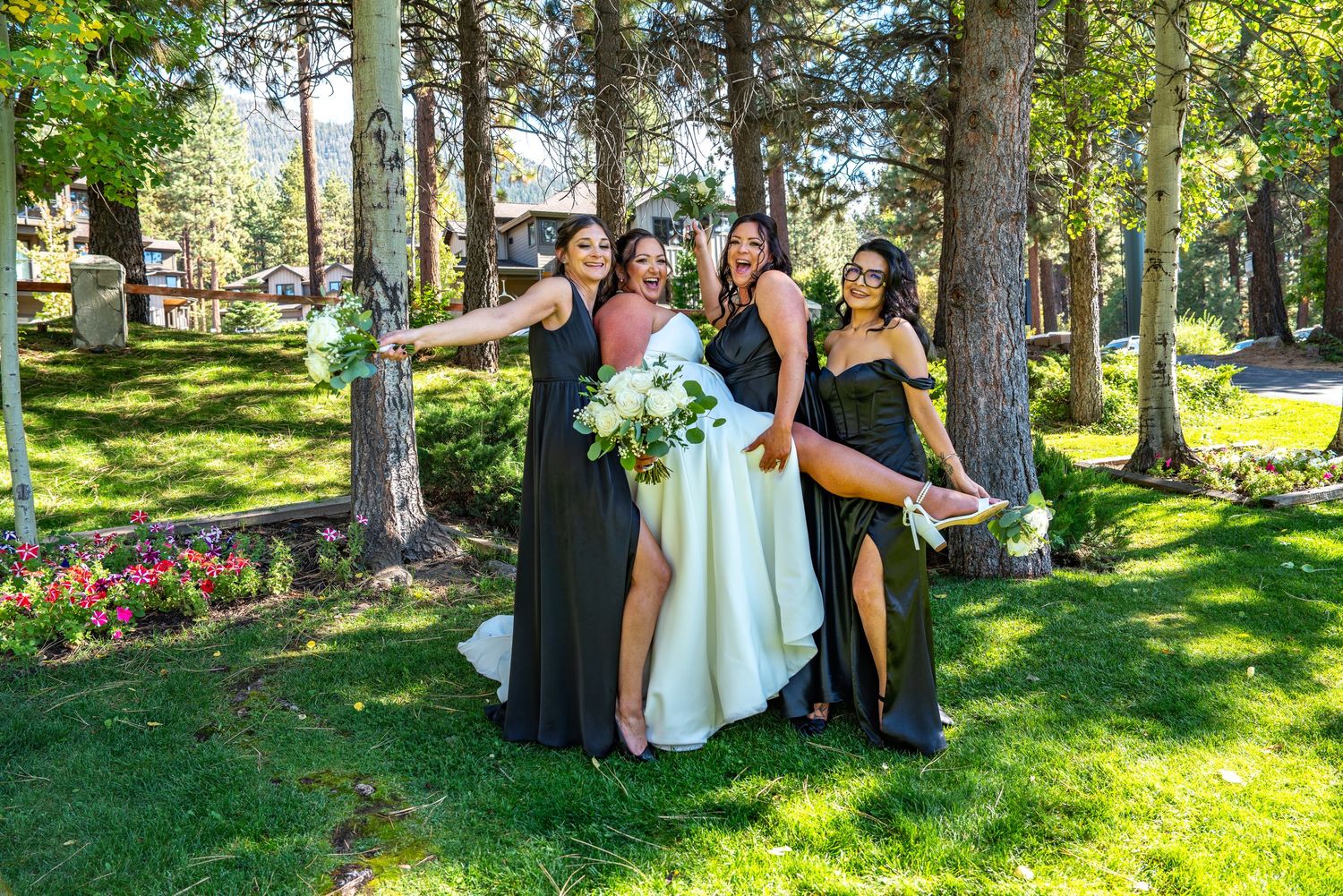 Bride and bridesmaids in dark green gowns pose joyfully outdoors among tall pine trees holding white bouquets.