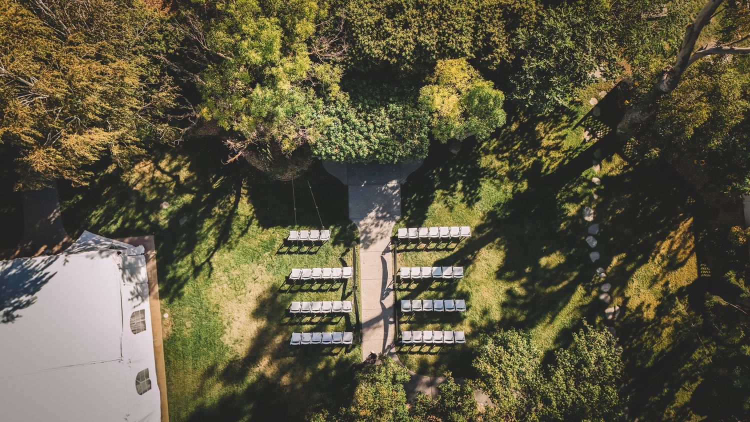 Aerial view of an outdoor wedding ceremony with white chairs arranged in rows amid lush green trees.