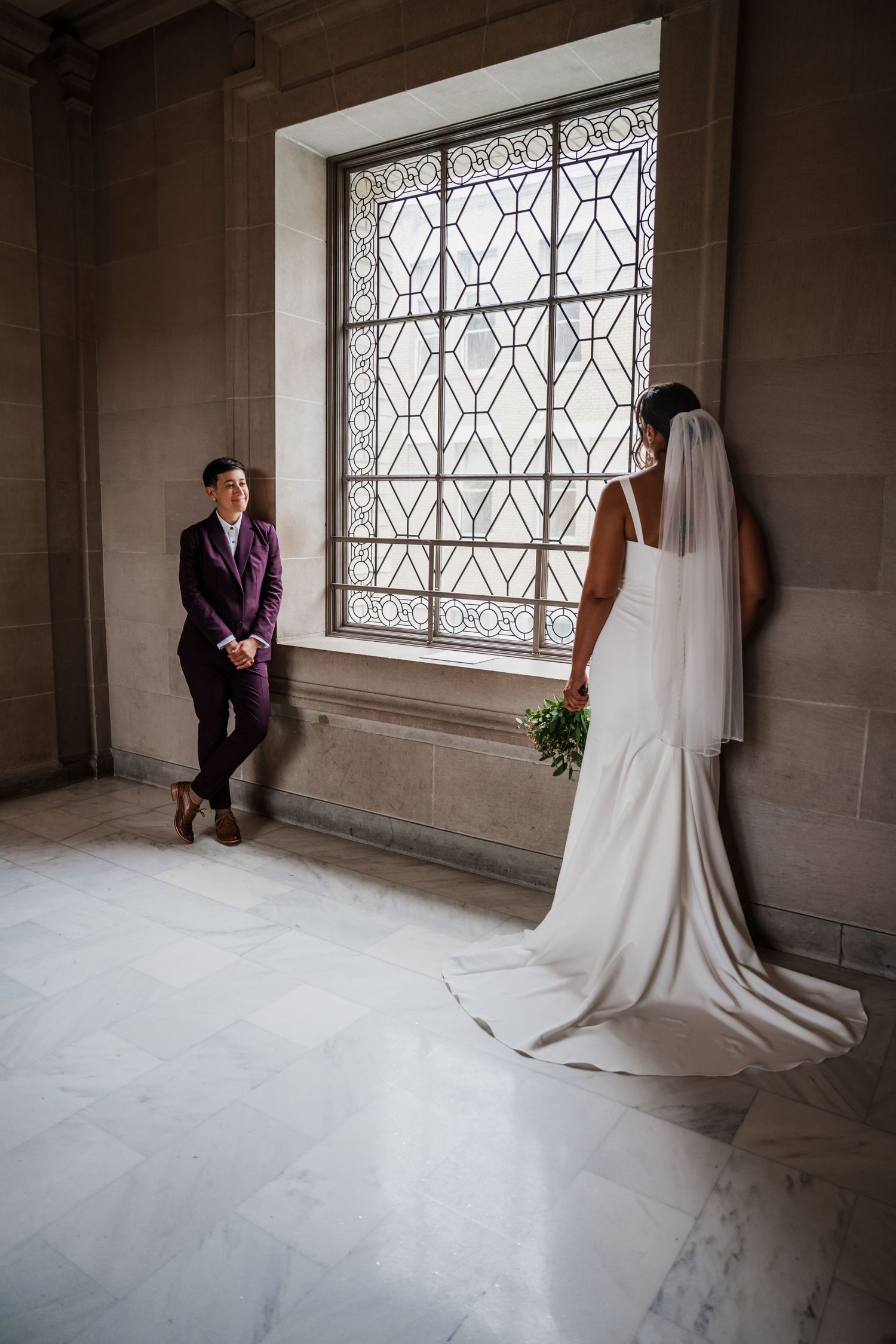 Wedding couple in elegant stone hallway with ornate windows creating dramatic lighting.