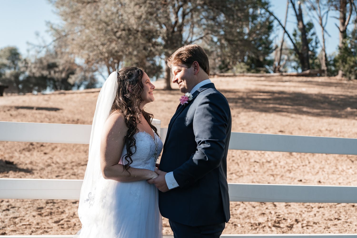 Bride in a white lace gown and veil holds hands with a groom in a navy suit beside a white ranch fence on a sunny outdoor wedding day.