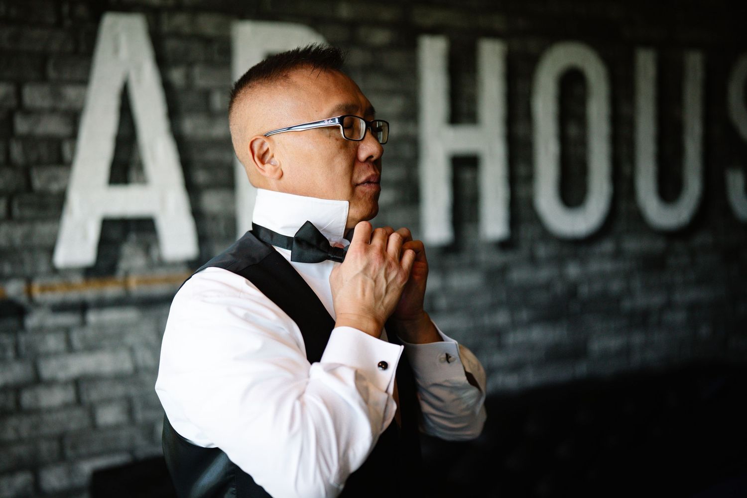 Man in white dress shirt, black vest and bow tie stands thoughtfully in front of a dark brick wall.