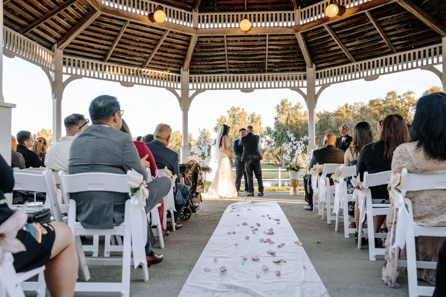 Outdoor wedding ceremony under a white gazebo with guests seated in white chairs along a floral aisle runner.
