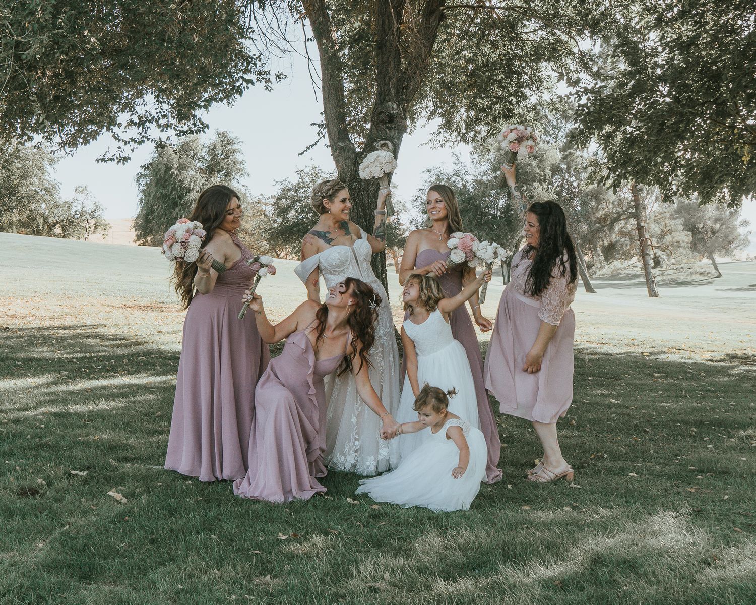 Bride surrounded by bridesmaids in lavender dresses posing under a tree at outdoor wedding.
