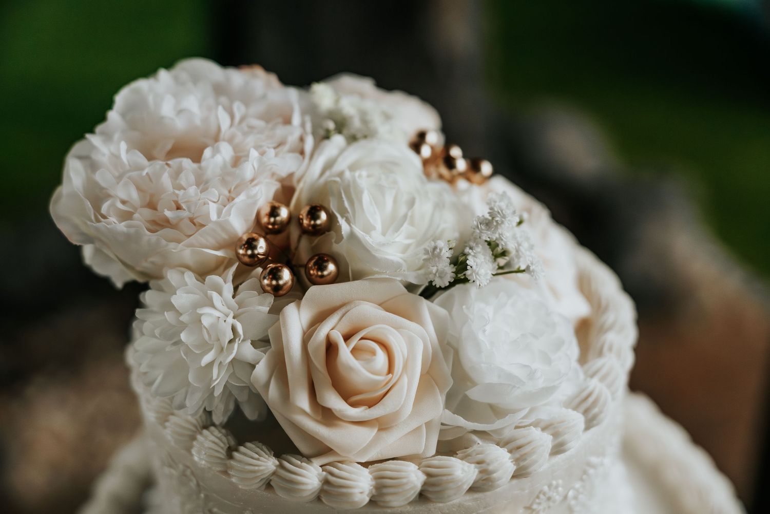 Elegant white wedding cake adorned with ivory roses, white flowers, and gold berry accents on a pearl border.