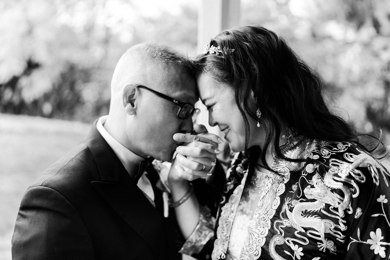 Black and white wedding photo of couple sharing tender moment, groom kissing bride's hand as she smiles joyfully.