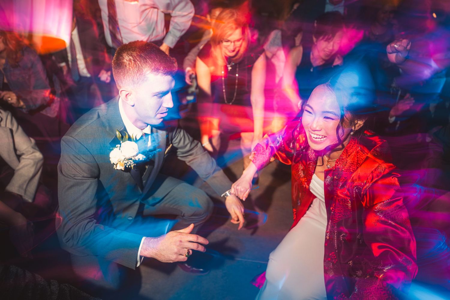 Bride in white gown and red jacket dances joyfully with groom at colorful wedding reception.