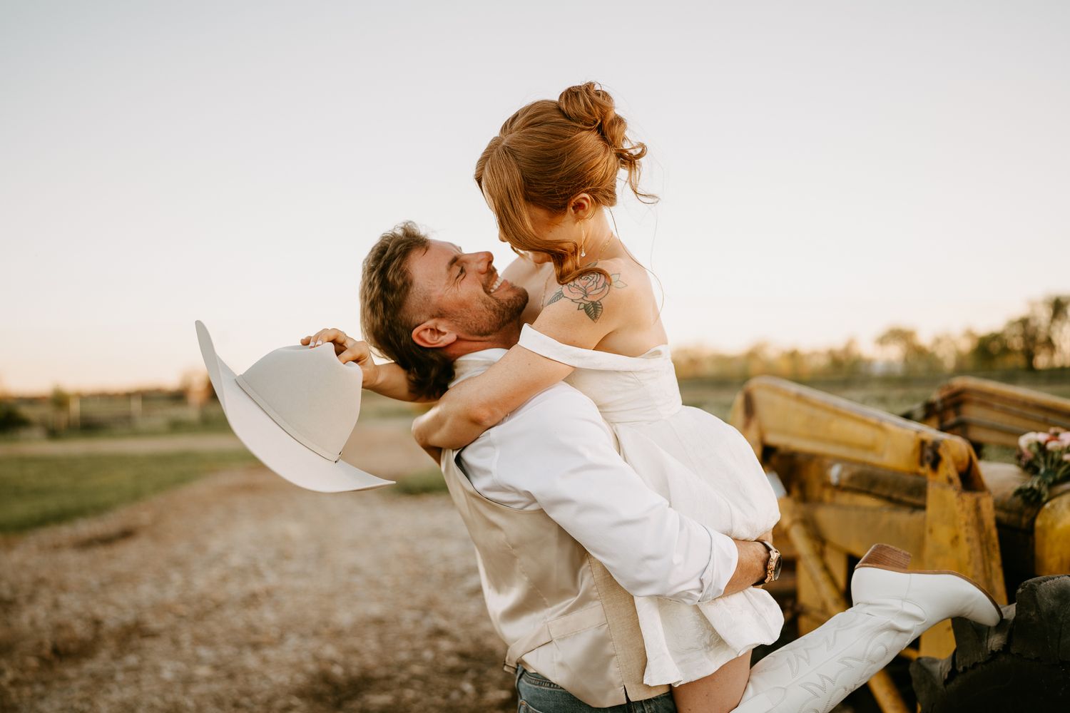 A groom lifts his bride in a sun-kissed outdoor wedding photo, sharing a romantic moment near a rustic wooden fence.