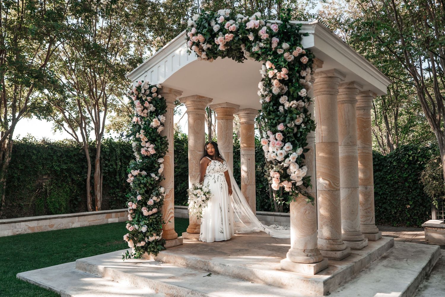 Bride in a flowing white gown and veil stands inside an elegant stone gazebo draped with lush pink and white floral garlands.