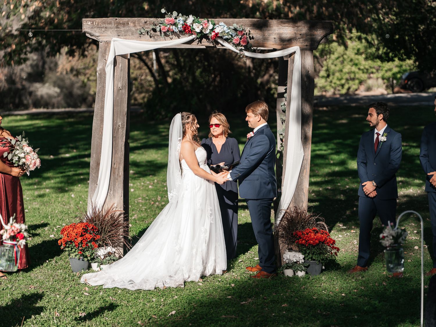 Bride in a white gown and groom in a navy suit exchange vows under a floral wooden arch during an outdoor garden wedding ceremony.