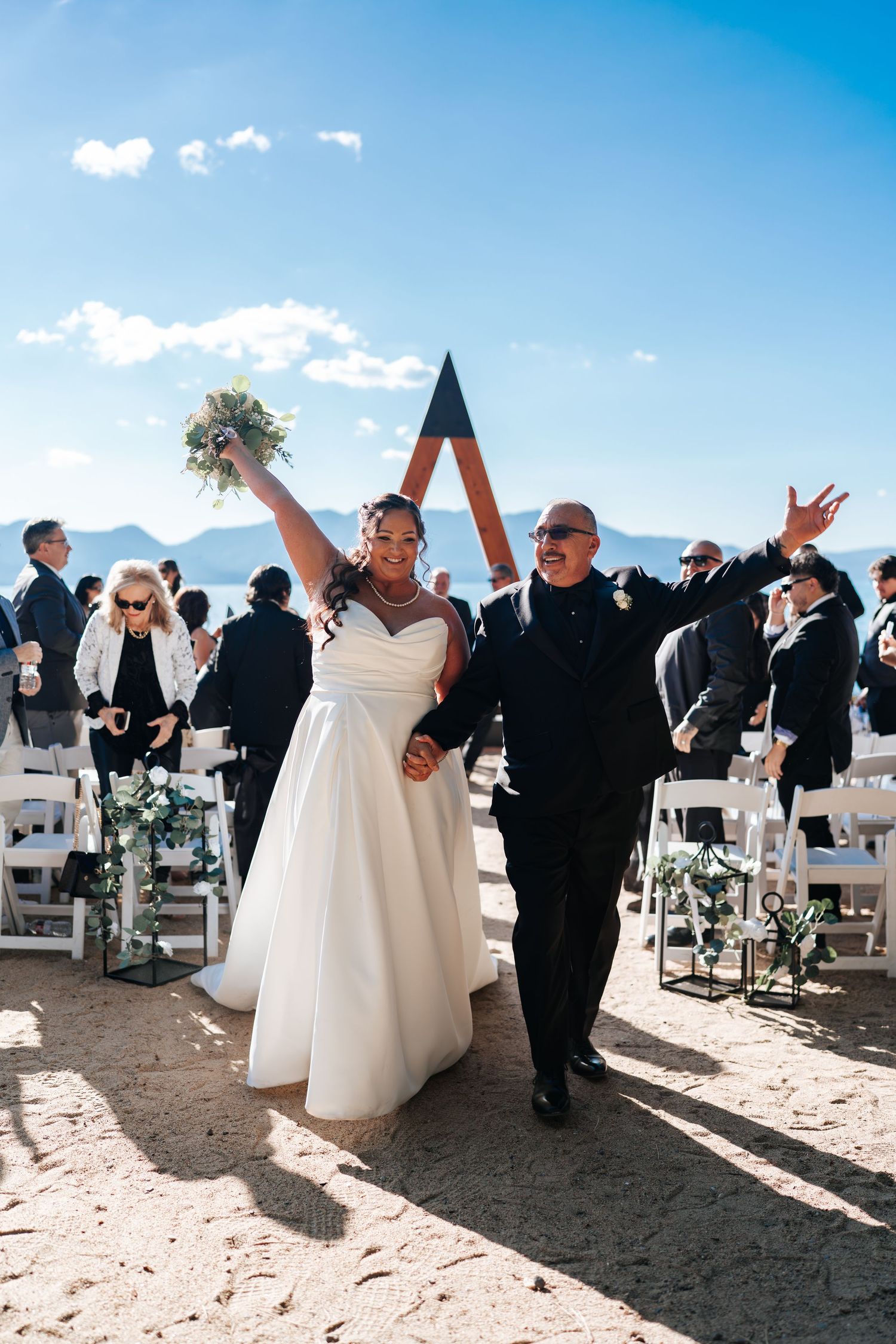 Beach wedding ceremony with guests seated in white chairs against blue sky.