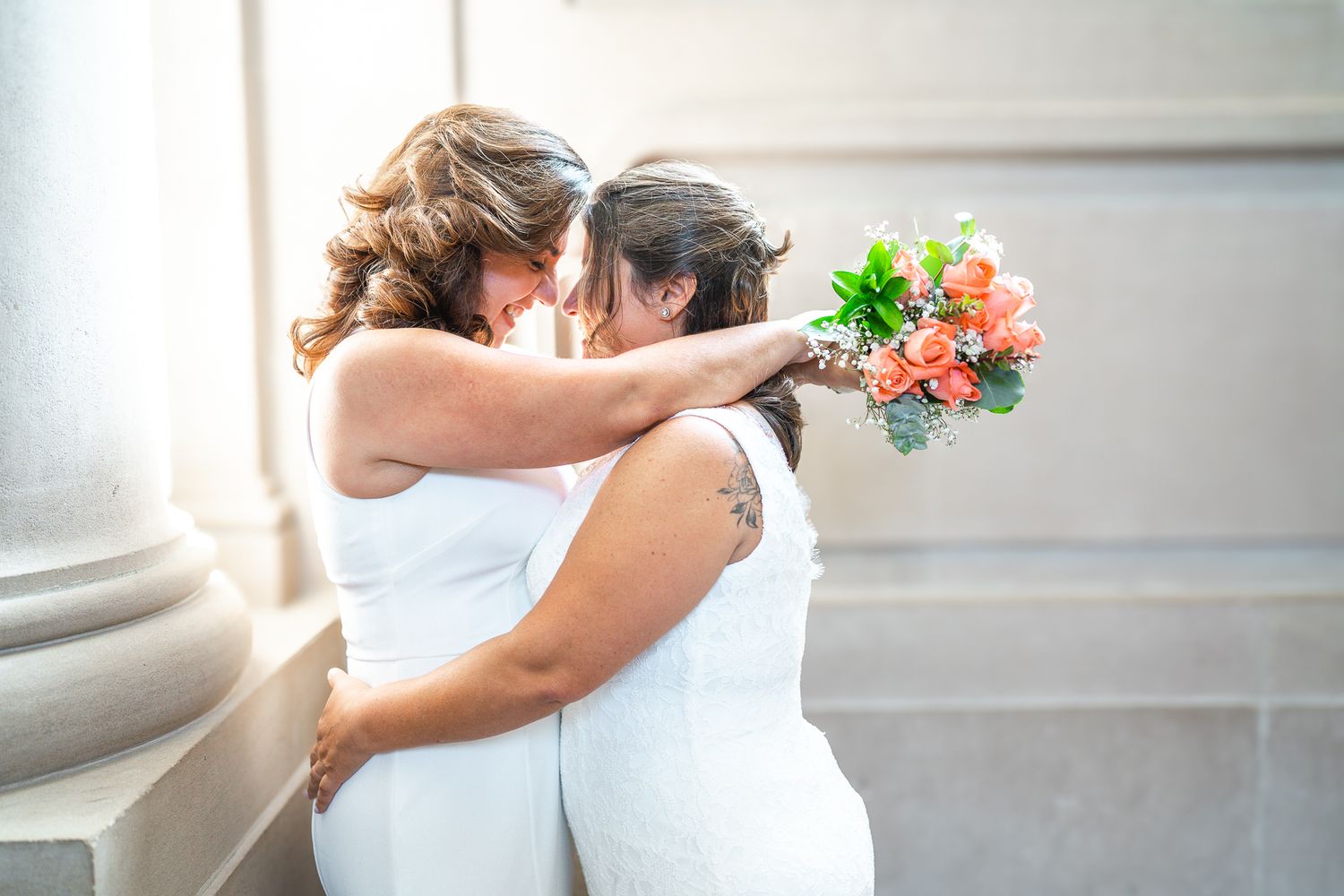Two brides in white dresses embrace tenderly, one holding a coral bouquet, in an outdoor wedding ceremony.