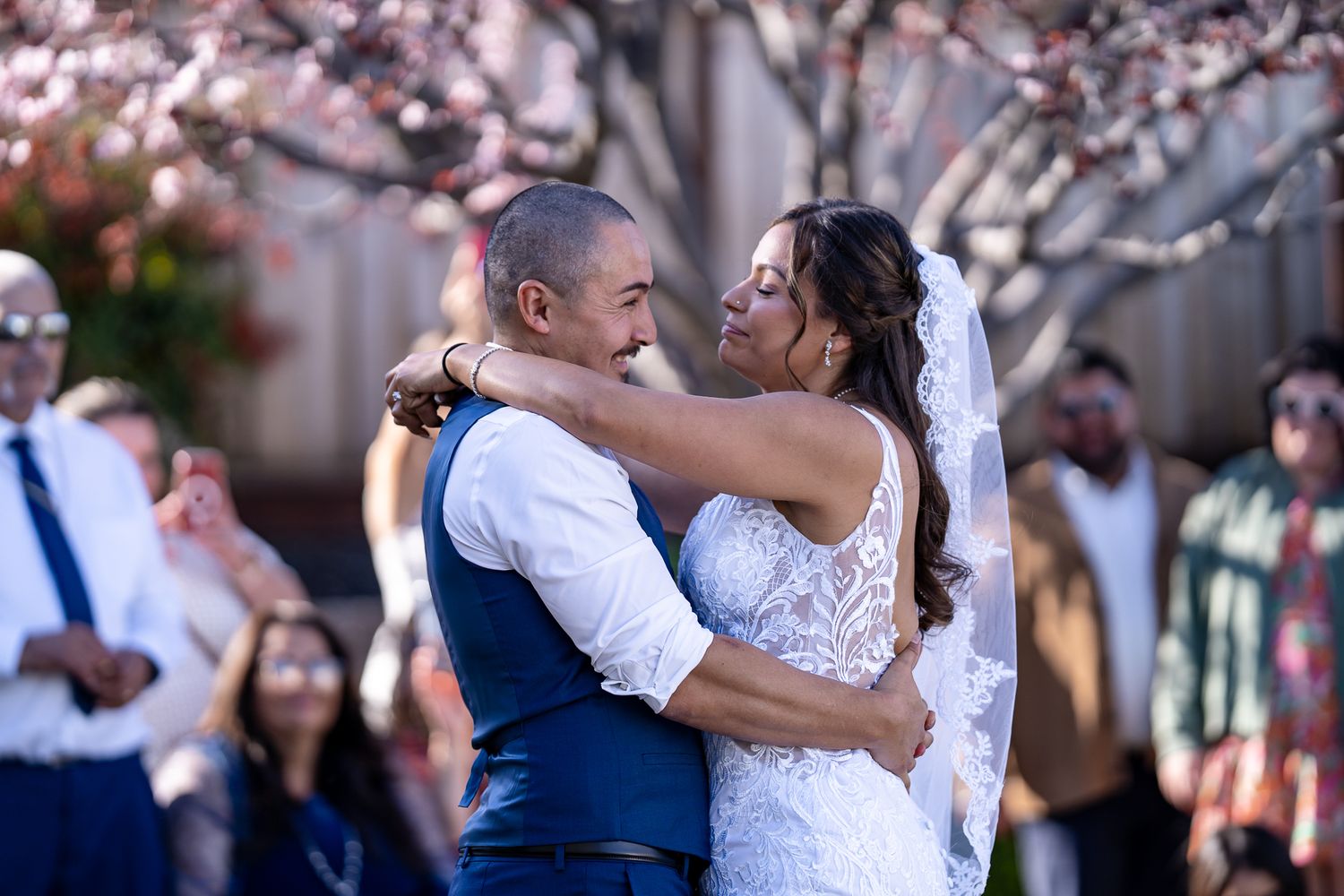 Bride and groom share first dance embrace outdoors under blooming cherry blossom trees at wedding ceremony.