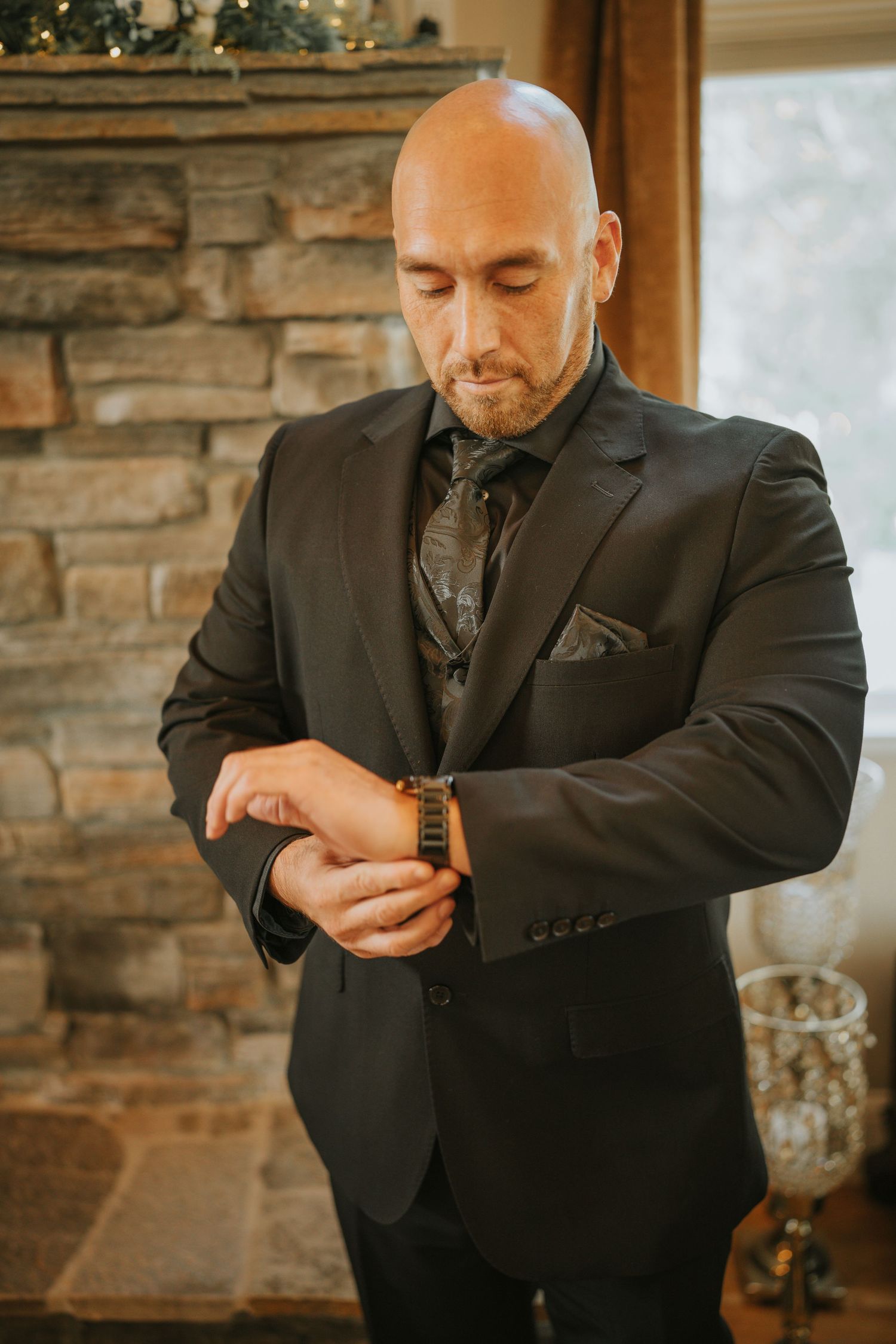 Bald man in black suit adjusting his wooden watch, standing in front of a stone fireplace.