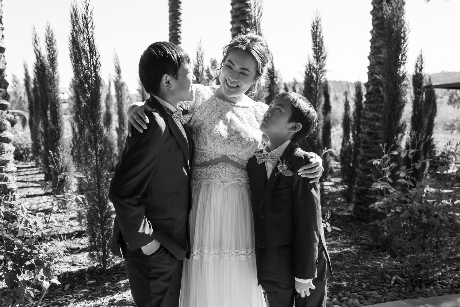 Bride in white gown smiling with two boys in suits outdoors among tall cypress trees in black and white photo.