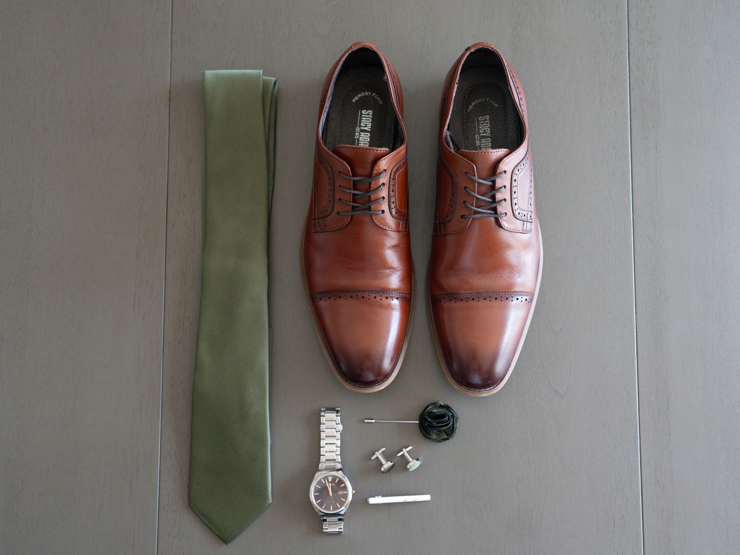Brown leather Oxford dress shoes displayed with olive green tie, silver watch, cufflinks, and accessories on gray background.