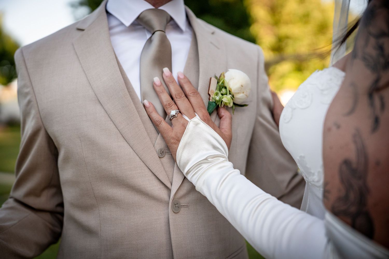 Bride's tattooed hand with ring resting on groom's beige suit with white boutonniere at wedding ceremony.