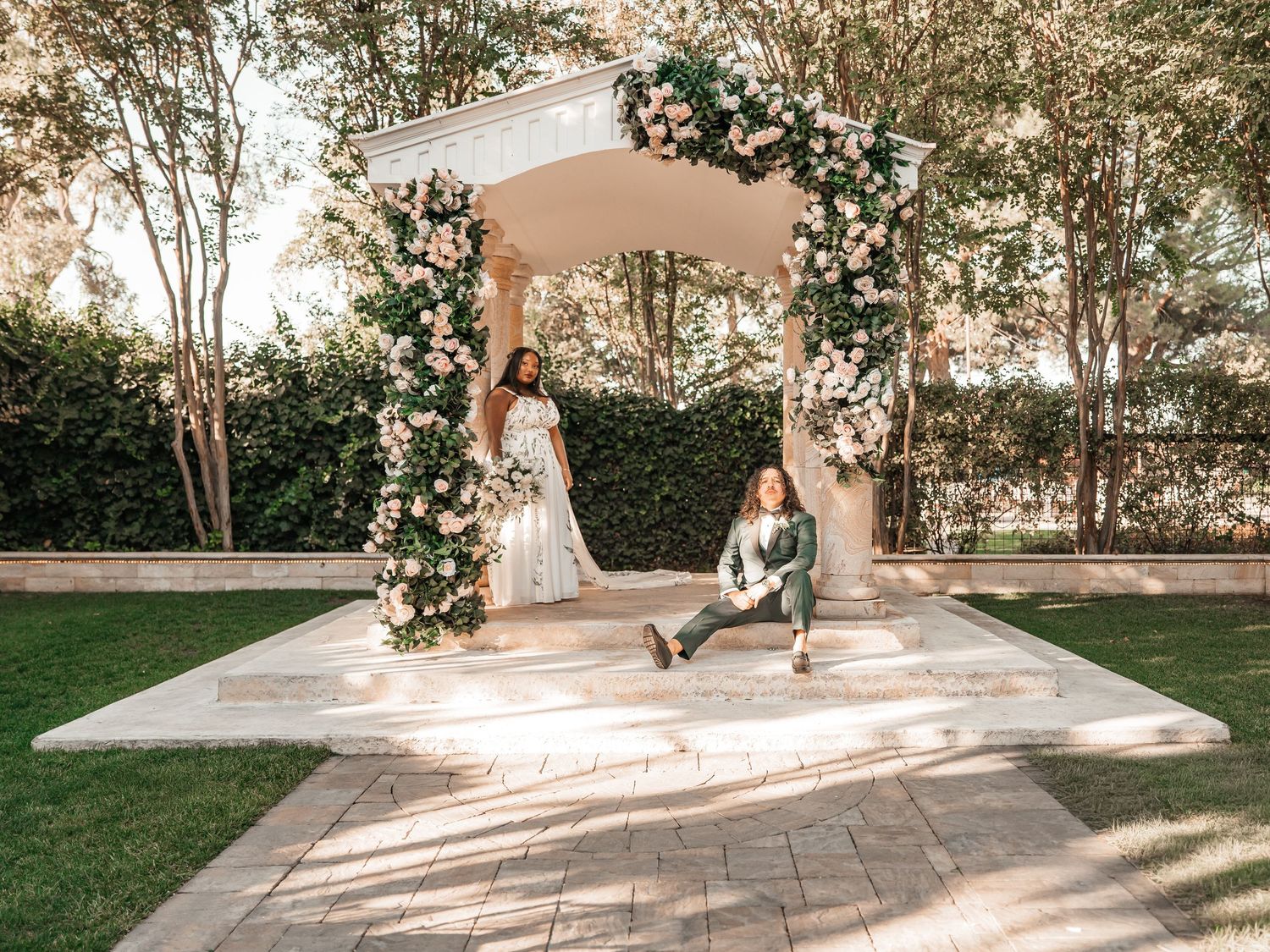 Bride in a white gown stands inside a floral-draped garden gazebo while a seated companion relaxes on the stone steps.