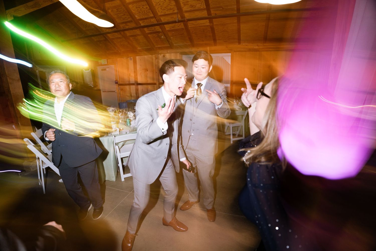 Guests dancing and singing at a lively wedding reception with colorful lights.