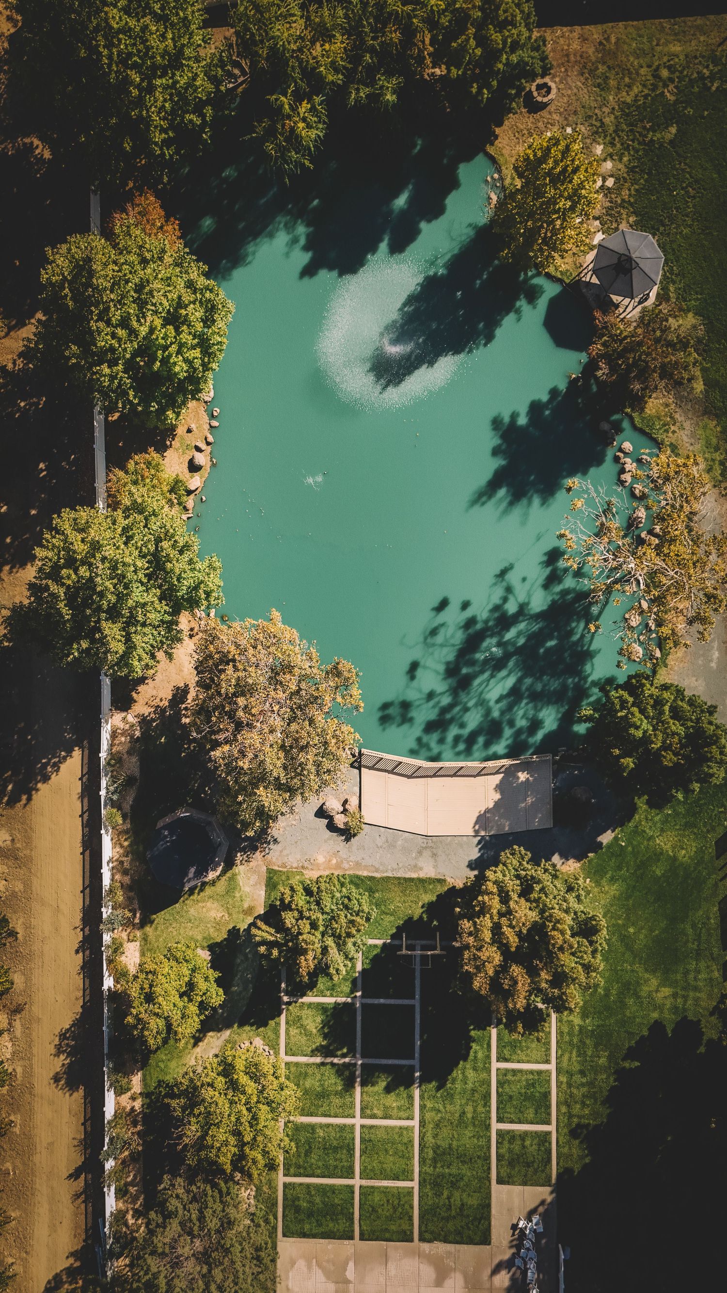 Aerial drone view of a turquoise pool surrounded by lush green trees casting shadows on a landscaped garden.