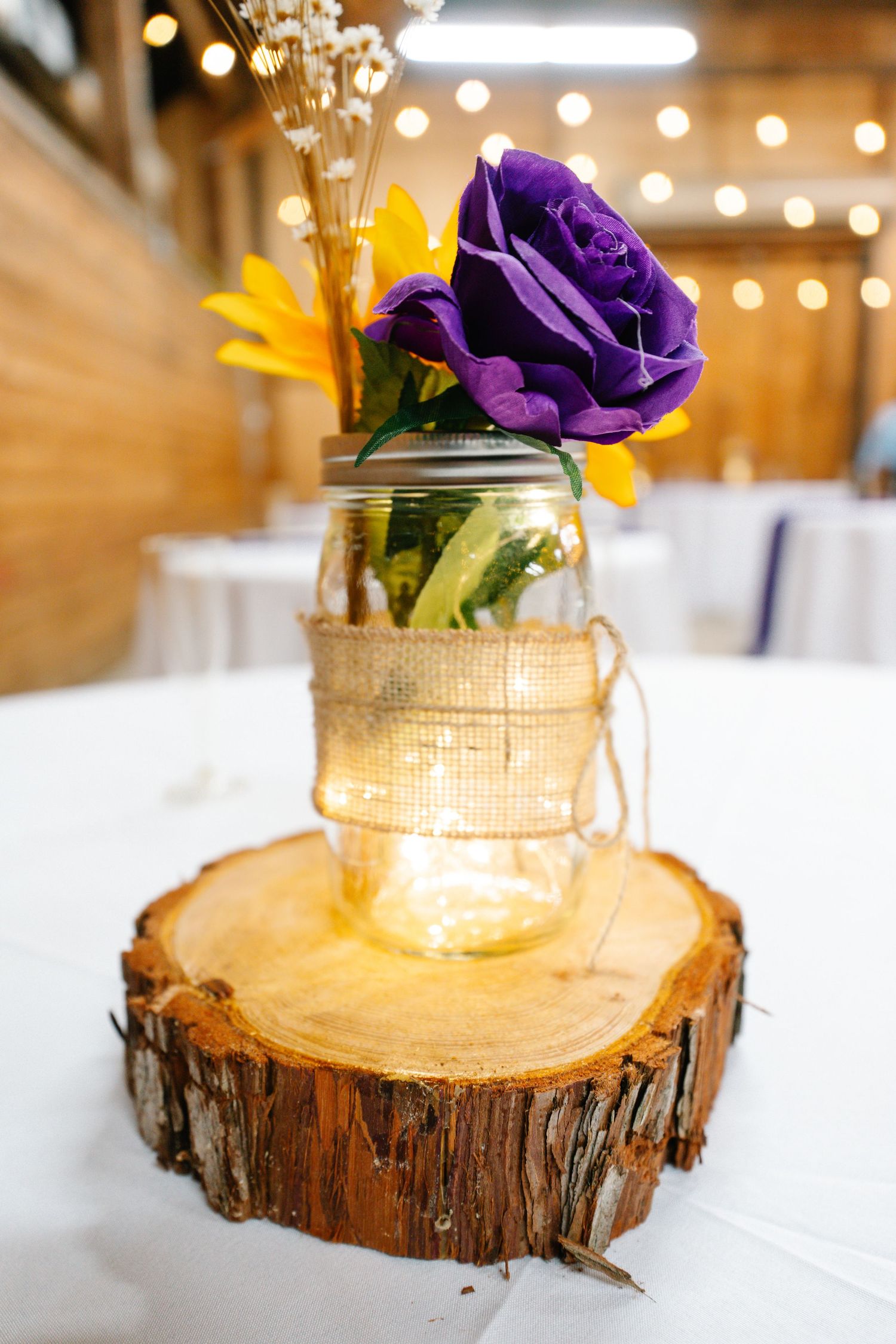 Rustic wedding centerpiece with purple rose and sunflowers in a mason jar on a wood slice.
