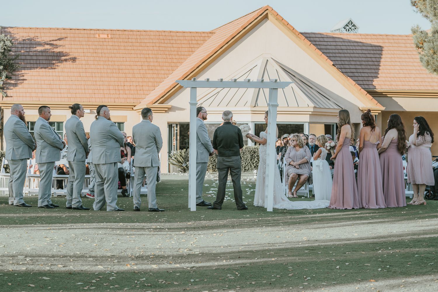 Wedding ceremony on a golf course with bridesmaids in pink and groomsmen in gray suits outdoors.