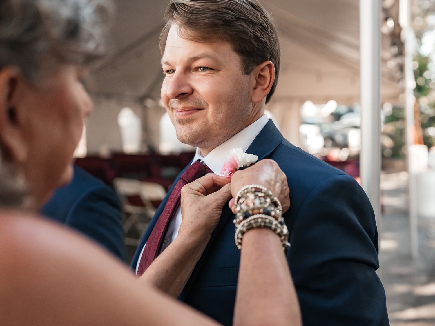 A woman with beaded bracelets pins a white boutonniere onto the navy suit lapel of a smiling man in a burgundy tie at an outdoor event.