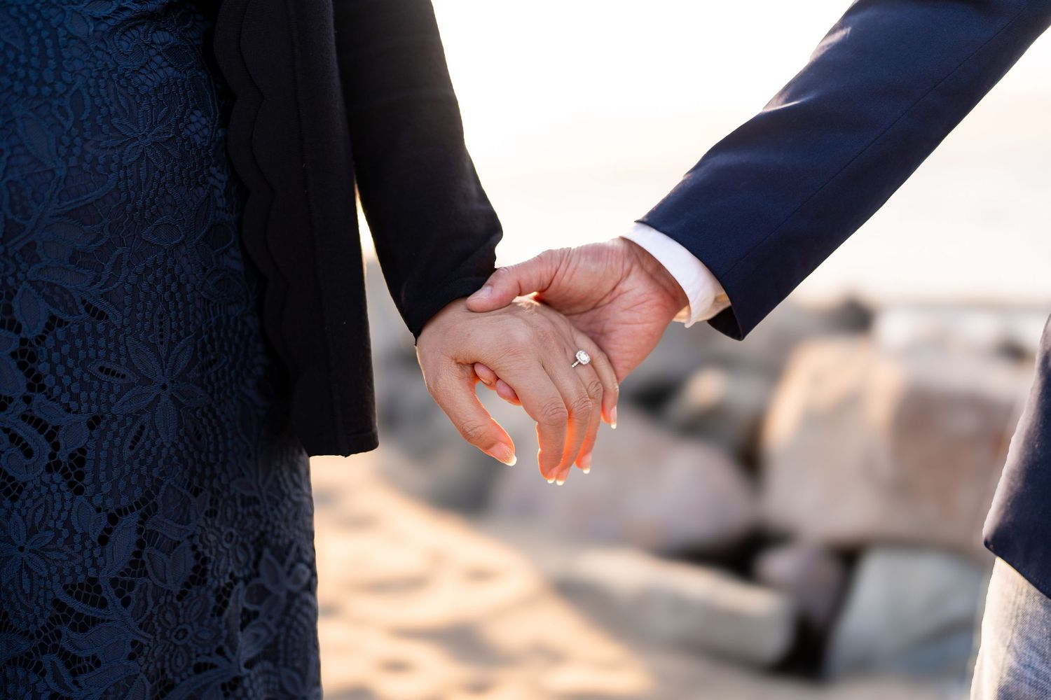 Couple walking hand in hand along rocky beach shoreline at sunset.